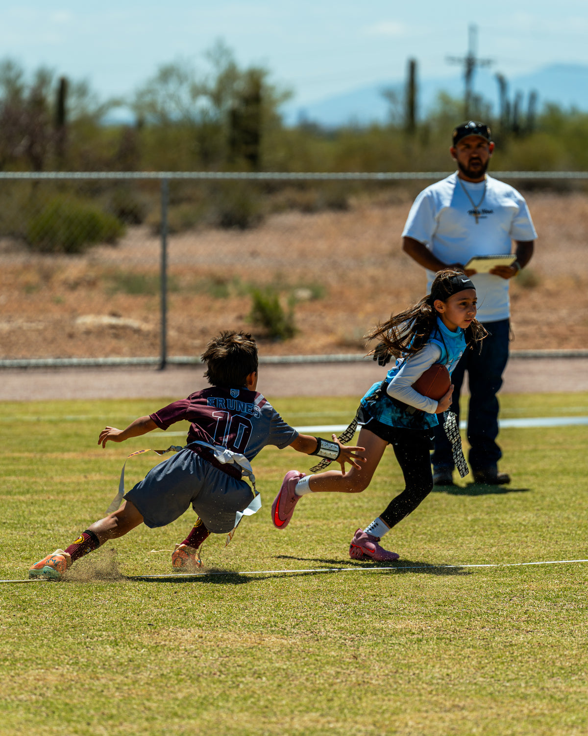 Two children playing flag football on a grassy field, with IN SZN Coach standing in the background near a desert landscape.
